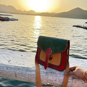 Puede incluir: Un bolso de cuero multicolor con solapa verde, centro marrón y ribete rojo. El bolso tiene una correa larga marrón y un cierre rojo. El fondo muestra una puesta de sol sobre un lago y montañas.