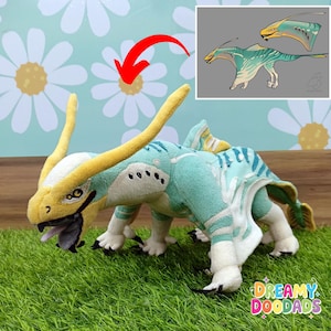 May include: A plush toy creature in shades of teal, yellow, and white, with a detailed design and a small, open mouth. The toy is on a bed of green faux grass, with a drawing of the creature in the upper right corner. The text "Dreamy Doodads" is visible.
