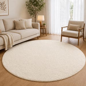 May include: A large, cream-coloured, circular rug is the focal point of a living room. The rug sits on a wooden floor, with a beige sofa, armchair, and a wooden side table with a lamp in the background. The room is bright with natural light.