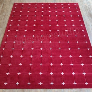 Op de afbeelding: Een groot, rechthoekig rood tapijt met een rasterpatroon van witte kruizen. Het tapijt is gemaakt van een zacht materiaal en is geschikt voor een woonkamer of slaapkamer. De afmetingen van het tapijt zijn niet zichtbaar.