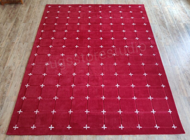 Op de afbeelding: Een groot, rechthoekig rood tapijt met een rasterpatroon van witte kruizen. Het tapijt is gemaakt van een zacht materiaal en is geschikt voor een woonkamer of slaapkamer. De afmetingen van het tapijt zijn niet zichtbaar.