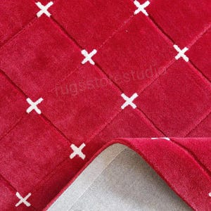 Op de afbeelding: Een close-up van een rode vloerkleed met een gewatteerd patroon en witte kruisaccenten. De textuur van het vloerkleed is zichtbaar en een hoek is teruggevouwen, waardoor een grijze achterkant zichtbaar is. Het vloerkleed is een woondecoratie-artikel.
