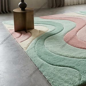 May include: A large, soft area rug with a wavy design in pastel colors. The rug features a pattern of flowing lines in shades of pink, mint green, and cream. A small, brown side table and a dark vase sit on the rug.