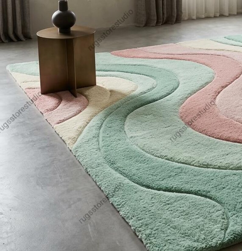 May include: A large, soft area rug with a wavy design in pastel colors. The rug features a pattern of flowing lines in shades of pink, mint green, and cream. A small, brown side table and a dark vase sit on the rug.