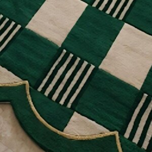 May include: A decorative rug with a scalloped edge. The rug features a green and white checkered pattern with alternating stripes. A gold-colored trim outlines the edge of the rug.