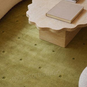 Peut inclure: Un tapis vert mousse présentant un motif répétitif de petits carrés en creux. Le tapis est placé sous une table basse en pierre beige à bords festonnés, sur laquelle reposent deux livres, dont un intitulé WORKSTEAD, et un canapé texturé blanc.