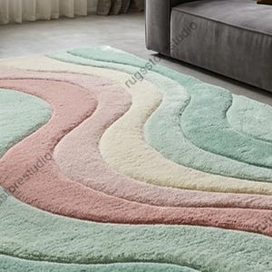 May include: A large, rectangular area rug with a wavy, layered design in pastel colors. The rug features soft, plush textures with bands of pink, green, cream, and off-white. The rug is placed on a light-colored floor, near a gray sofa.