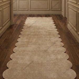 Peut inclure: Un tapis de couloir beige à bords festonnés. Le tapis est rectangulaire avec une bordure ondulée et est posé sur un plancher en bois foncé. Les murs sont de couleur beige clair avec des panneaux décoratifs. Le tapis ajoute une touche d'élégance.