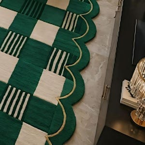 May include: A green and cream-colored area rug with a unique scalloped edge. The rug features a checkerboard pattern with alternating green squares and cream squares, some with white stripes. A gold trim outlines the edge.