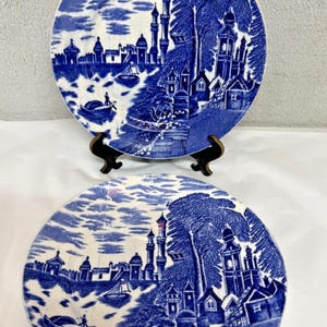 May include: Two round ceramic plates featuring a detailed blue and white transferware pattern. The design depicts a coastal landscape with tall towers, buildings, trees, and small boats on the water. The plates are displayed on a black stand.