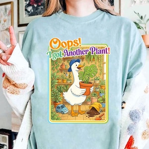 Puede incluir: Camiseta azul claro con un dibujo animado de un ganso con un sombrero azul, sosteniendo una maceta. El gráfico incluye el texto "Oops! I got Another Plant!" en naranja y verde. El diseño presenta un coche azul pequeño y varias plantas en macetas.