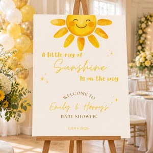 May include: A rectangular white baby shower welcome sign on a wooden easel featuring a yellow watercolor sun illustration and the text: A little ray of Sunshine is on the way, WELCOME TO Emily & Harry's BABY SHOWER, 5 JULY 2026.