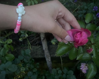 May include: A bracelet with a white turtle charm, white beads, and pink and white disc beads. The bracelet is worn on a wrist near a pink rose in a garden. The bracelet is a charming accessory.