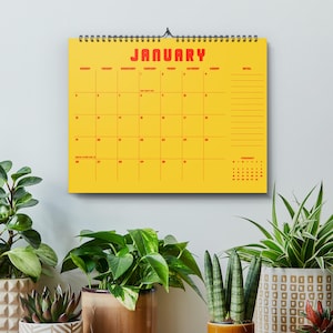 Könnte beinhalten: Ein gelber Wandkalender für Januar mit rotem Text und Rasterlinien, der über einer Holzoberfläche hängt. Der Kalender ist von verschiedenen Topfpflanzen mit grünem Laub umgeben.