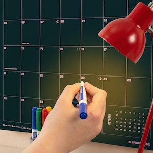 Puede incluir: Un calendario de pared verde oscuro con dise&ntilde;o de cuadr&iacute;cula, l&iacute;neas y n&uacute;meros rosas para el mes de octubre. Una mano sostiene un marcador borrable azul cerca de la fecha 23. Una l&aacute;mpara de escritorio roja brillante ilumina la superficie.