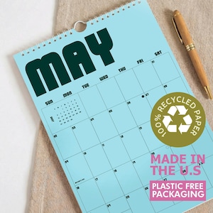 Puede incluir: Un calendario de pared azul claro para el mes de mayo con letras oscuras en negrita. El calendario presenta un dise&ntilde;o de cuadr&iacute;cula con los d&iacute;as de la semana, un peque&ntilde;o calendario de referencia de junio y una pegatina dorada 100% RECYCLED PAPER.