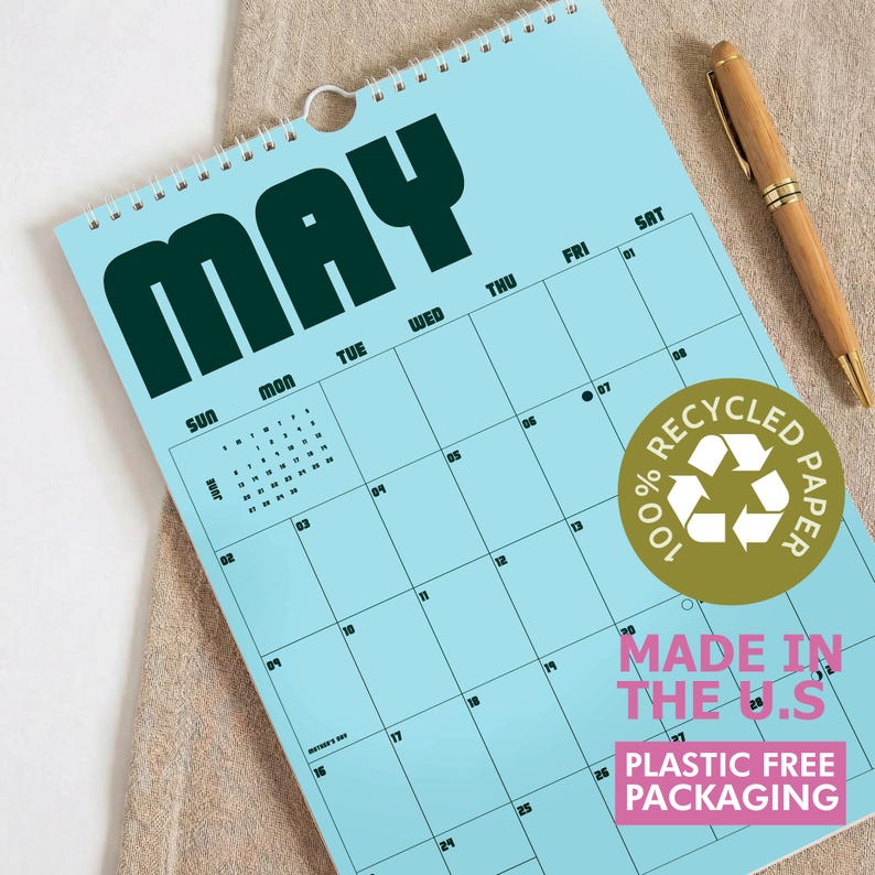 Puede incluir: Un calendario de pared azul claro para el mes de mayo con letras oscuras en negrita. El calendario presenta un dise&ntilde;o de cuadr&iacute;cula con los d&iacute;as de la semana, un peque&ntilde;o calendario de referencia de junio y una pegatina dorada 100% RECYCLED PAPER.