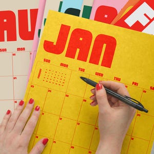 Puede incluir: Una p&aacute;gina de calendario de pared amarilla para enero con texto rojo en negrita y un dise&ntilde;o de cuadr&iacute;cula. El calendario presenta una peque&ntilde;a cuadr&iacute;cula de referencia de febrero en la esquina superior izquierda. Un bol&iacute;grafo negro est&aacute; sobre la cuadr&iacute;cula.