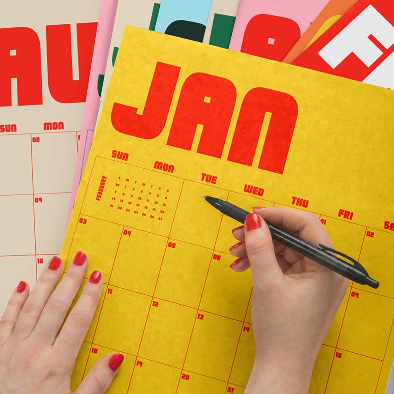 Puede incluir: Una p&aacute;gina de calendario de pared amarilla para enero con texto rojo en negrita y un dise&ntilde;o de cuadr&iacute;cula. El calendario presenta una peque&ntilde;a cuadr&iacute;cula de referencia de febrero en la esquina superior izquierda. Un bol&iacute;grafo negro est&aacute; sobre la cuadr&iacute;cula.