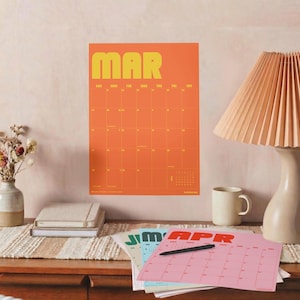 Puede incluir: Un calendario de pared para marzo con un fondo naranja brillante y texto amarillo que dice MAR. Debajo, una cuadr&iacute;cula muestra los d&iacute;as del mes. Un segundo calendario para abril con fondo rosa y texto rojo APR sobre el escritorio.