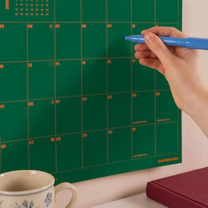 Puede incluir: Un calendario de pared verde oscuro con l&iacute;neas de cuadr&iacute;cula y n&uacute;meros de fecha en naranja. El calendario incluye texto con nombres de meses y etiquetas de festividades como Christmas Eve y New Year's Eve. Un marcador azul se sostiene.