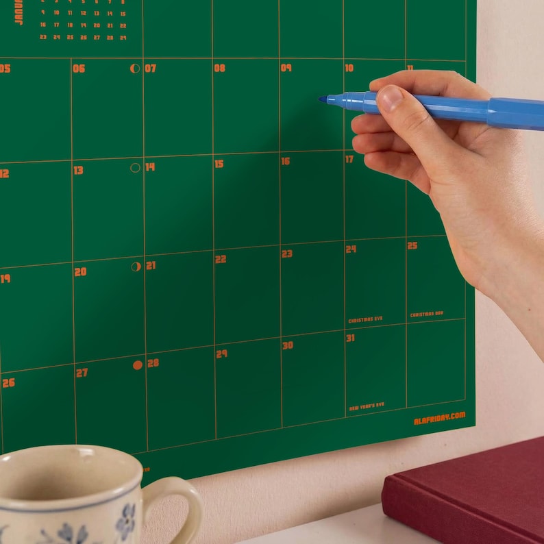 Puede incluir: Un calendario de pared verde oscuro con l&iacute;neas de cuadr&iacute;cula y n&uacute;meros de fecha en naranja. El calendario incluye texto con nombres de meses y etiquetas de festividades como Christmas Eve y New Year's Eve. Un marcador azul se sostiene.