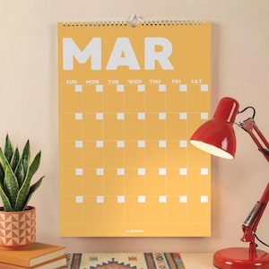 Peut inclure: Un calendrier mural jaune pour le mois de mars avec du texte blanc et une grille de carrés. Une lampe de bureau rouge est à droite et une plante en pot à gauche. Le calendrier est accroché au mur.