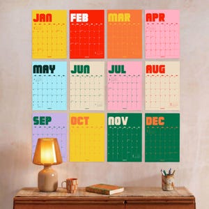May include: A wall calendar set featuring twelve individual monthly sheets in vibrant colors including yellow, red, orange, pink, blue, green, and purple. Each sheet displays the month name in bold letters and a grid for daily planning.