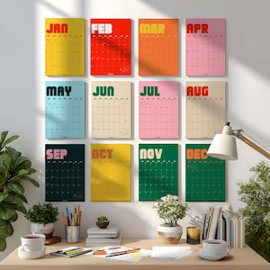 Könnte beinhalten: Ein Set aus zwölf Wandkalendern, die in einem Raster angeordnet sind. Jeder Kalender repräsentiert einen Monat von Januar bis Dezember mit individuellen Farbschemata und fetter Typografie. Die Kalender zeigen ein Datumsraster an der Wand.