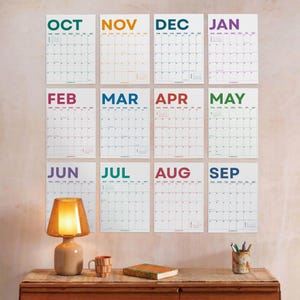 May include: Twelve individual wall calendar sheets arranged in a grid, each featuring a different month with bold, colorful headers. Below the calendar display sits a warm-toned amber glass lamp, a striped ceramic mug, a book, and a pen holder.
