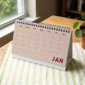 May include: A desktop spiral-bound calendar for the month of January, featuring a light beige background with dark red text for the days of the week and the month name JAN. The grid displays dates from 1 to 31 with lines for notes at the bottom.