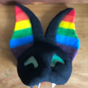 May include: A plush bat head with black fabric and large, upright ears featuring horizontal stripes in red, orange, yellow, green, blue, and purple. The face has two light green, triangular eye cutouts and two small, white, pointed fangs protruding from the bottom.