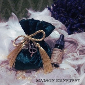 May include: A dark green velvet pouch tied with a gold rope and tassels, adorned with a silver four-leaf clover charm. A small bottle with a dropper and a label is next to the pouch. The text "MAISON ERNSTSSY" is at the bottom.