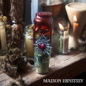Könnte beinhalten: Ein Glasgefäß gefüllt mit Kräutern und einem roten Herzanhänger. Das Glas ist mit dunkelrotem Wachs versiegelt und trägt unten den Text "MAISON ERNSTSSY". Weitere Kerzen und Gläser befinden sich im Hintergrund.