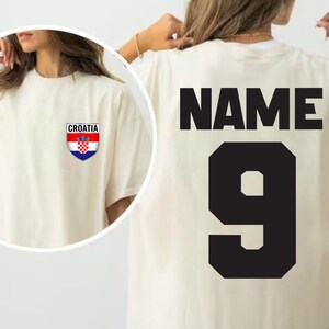 May include: Cream-colored oversized t-shirt with the word "NAME" and the number "9" in black on the back. A small Croatian flag emblem is on the front. The shirt has short sleeves and a relaxed fit.