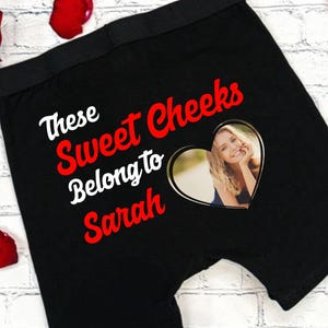 May include: Black boxer shorts with the text "These Sweet Cheeks Belong to Sarah" in white and red. A heart-shaped photo of a woman is featured on the front. Red rose petals surround the shorts.