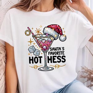 Op de afbeelding: Wit T-shirt met een feestelijk cocktailontwerp. Het ontwerp bevat een kerstmuts, ijsblokjes en de tekst "Santa's Favorite Hot Mess". Er zijn ook gouden accenten en sterren. De onderkant van de afbeelding bevat de tekst "CHRISTMAS HOODIES JUMPERS AVAILABLE."
