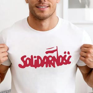 May include: White t-shirt with the Polish word "Solidarność" in red, featuring a stylized flag design. The shirt is made of a soft material and is suitable for casual wear.