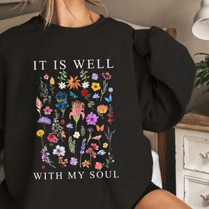 May include: Black sweatshirt featuring the phrase "IT IS WELL WITH MY SOUL" and a vibrant floral arrangement. The design incorporates diverse flowers, butterflies, and foliage in pink, yellow, blue, and purple hues.