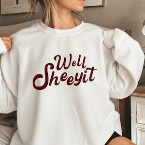 May include: A white long-sleeve crewneck sweatshirt featuring the dark red text "Well Sheeyit" printed in a stylised cursive font across the chest. The garment has a relaxed fit with ribbed cuffs and a ribbed neckline, made of a solid white fabric.