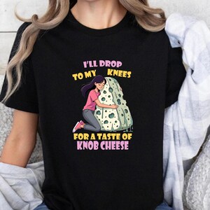 Puede incluir: Camiseta negra con un dibujo de una persona abrazando un gran trozo de queso. El texto dice: "I'll drop to my knees for a taste of knob cheese" en rosa y amarillo.
