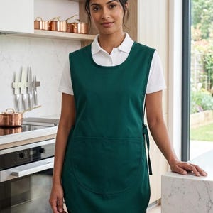 May include: A dark green tabard-style apron featuring a rounded neckline, side ties for an adjustable fit, and a large, deep front pocket. The apron is worn over a white collared polo shirt and black pants, providing full front coverage.