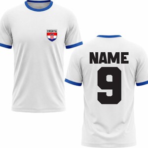 Puede incluir: Camiseta de f&uacute;tbol blanca con ribetes azules en el cuello y las mangas. La parte delantera presenta un escudo croata y la palabra "CROATIA". La parte trasera muestra la palabra "NAME" sobre el n&uacute;mero "9" en negro.
