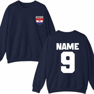 May include: Navy blue sweatshirt with a Croatia crest on the front. The back of the sweatshirt displays the word "NAME" above the number "9" in white block letters. A sporty apparel item.