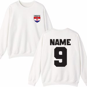 May include: White sweatshirt with the Croatian flag emblem on the front. The back of the sweatshirt features the word "NAME" above the number "9" in black block letters. A casual, sporty design.