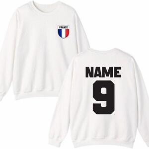 May include: White sweatshirt with a French flag emblem on the chest and the word "FRANCE" above it. The back of the sweatshirt features the word "NAME" above the number "9" in black. A casual, sporty design.