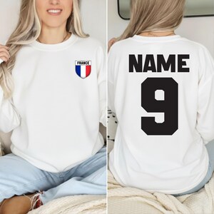 May include: White sweatshirt with a small France emblem on the front and the word "NAME" and the number "9" on the back in black. The France emblem is a shield with blue, white, and red vertical stripes.