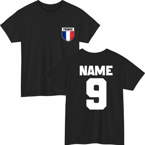 May include: Black t-shirt with a French flag emblem on the chest. The back of the shirt displays the word "NAME" above the number "9" in white block letters. A sporty design.