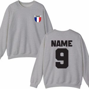 May include: A heather gray sweatshirt with a crew neck. The front features a small patch with the word "FRANCE" and a blue, white, and red shield design. The back displays the word "NAME" above the number "9" in black.