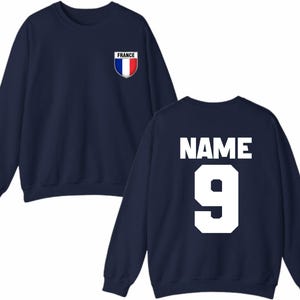 May include: Navy blue sweatshirt with the French flag emblem on the chest. The back of the sweatshirt displays the word "NAME" above the number "9" in white block letters. A casual, sporty design.